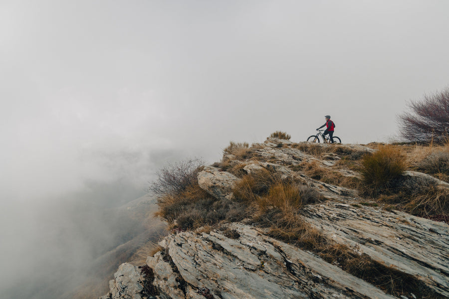 A rider standing over a mountain bike on a rocky ridge above the clouds.