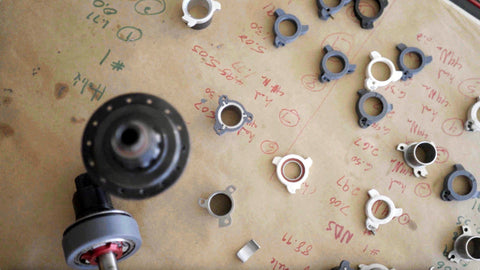 Dozens of 3D printed pusher prototypes laid out on a workbench covered in handwritten measurements, with a Sidekick hub and axle in the foreground.